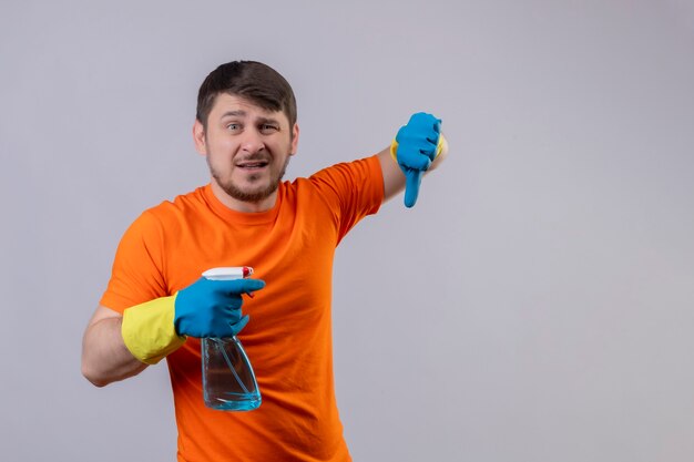 Junger Mann, der orange T-Shirt und Gummihandschuhe trägt, die Reinigungsspray mit traurigem Ausdruck halten