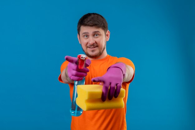 Junger Mann, der orange T-Shirt und Gummihandschuhe trägt, die Reinigungsspray und Schwamm halten