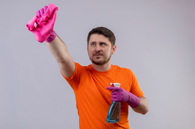 Junger Mann, der orange T-Shirt und Gummihandschuhe trägt, die Reinigungsspray und Teppich halten, die es betrachten