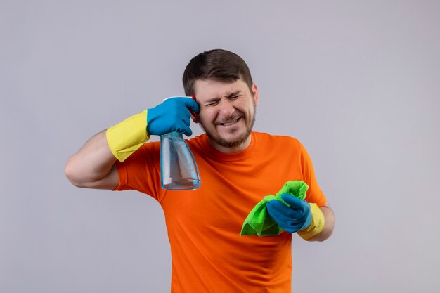 Junger Mann, der orange T-Shirt und Gummihandschuhe trägt, die Reinigungsspray und Teppich halten