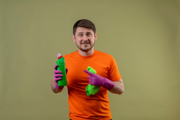 Junger Mann, der orange T-Shirt und Gummihandschuhe trägt, die Reinigungsspray und Teppich zeigen, der mit Finger zur Flasche mit Spray zeigt, der fröhlich positiv und glücklich betrachtet Kamera auf grünem Hintergrund lächelt