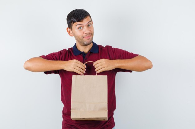 Junger mann, der papiertüte im t-shirt hält und fröhlich aussieht, vorderansicht.