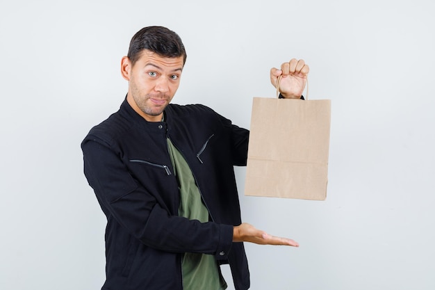 Junger Mann, der Papiertüte im T-Shirt, in der Jacke zeigt und fröhlich, Vorderansicht schaut.