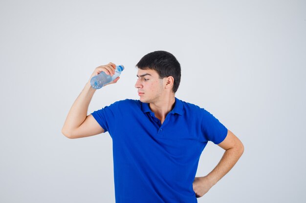 Junger Mann, der Plastikflasche hält, die Hand auf Taille im T-Shirt hält und zuversichtlich schaut. Vorderansicht.