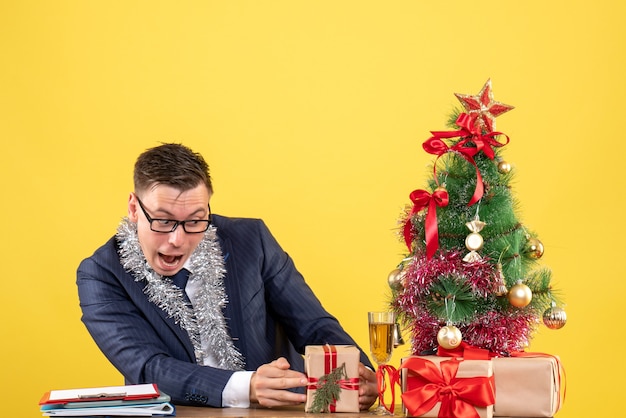 junger Mann, der sein Geschenk betrachtet, das am Tisch nahe Weihnachtsbaum sitzt und auf Gelb präsentiert