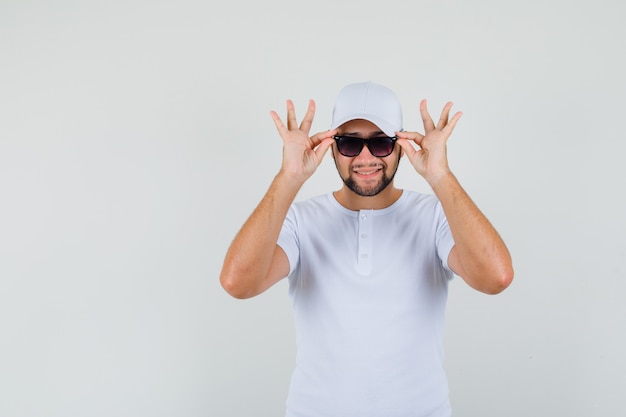 Junger Mann, der seine Brille berührt, während er in T-Shirt, Mütze lächelt und fröhlich aussieht. Vorderansicht.