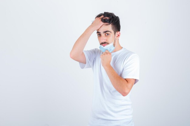 Junger Mann, der seine Maske abnimmt, die Haare mit der Hand im weißen T-Shirt kämmt und selbstbewusst aussieht