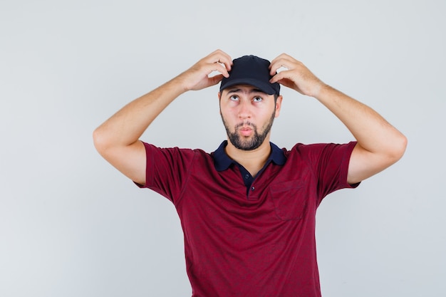 Junger Mann, der seine Mütze berührt, während er in rotem T-Shirt, Mütze und nachdenklichem Blick nach vorne schaut.