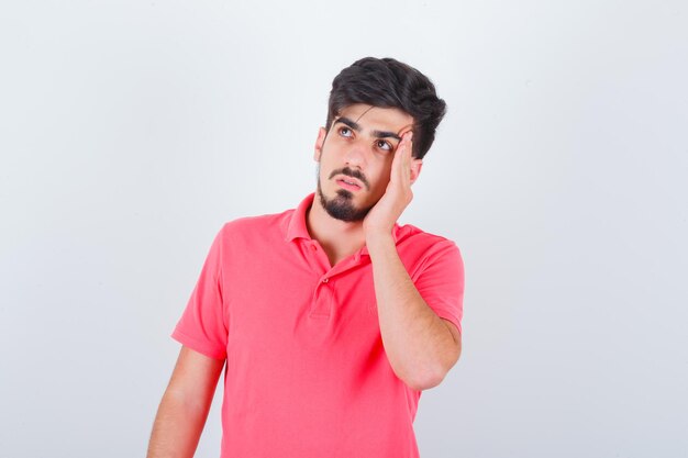 Junger Mann, der seine Schläfen im rosa T-Shirt reibt und nachdenklich aussieht. Vorderansicht.