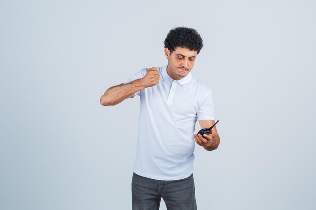 Junger Mann, der sich bereit macht, auf Walkie-Talkie-Telefon in weißem T-Shirt, Hose und gereiztem Blick zu schlagen, Vorderansicht.