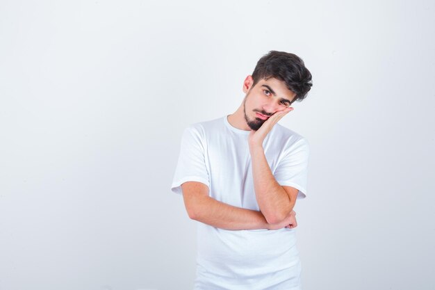 Junger Mann, der sich im T-Shirt die Wange zur Hand lehnt und wehmütig aussieht