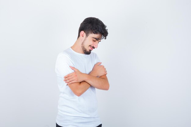 Junger Mann, der sich umarmt, während er im weißen T-Shirt nach unten schaut und hoffnungsvoll aussieht