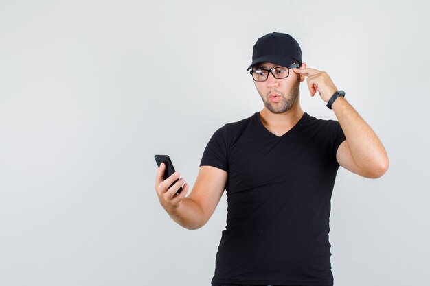Junger Mann, der Smartphone mit Finger auf Schläfen im schwarzen T-Shirt betrachtet