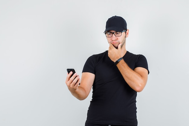 Junger Mann, der Smartphone mit Hand auf Kinn im schwarzen T-Shirt betrachtet