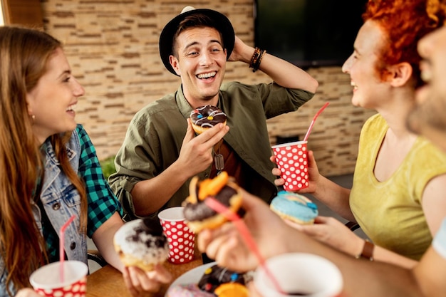 Junger Mann, der Spaß hat, während er Donut isst und mit seinen Freunden in einem Café spricht