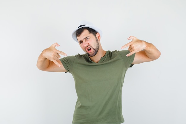 Junger Mann, der Steinsymbol im grünen T-Shirt und im Hut tut und cool schaut