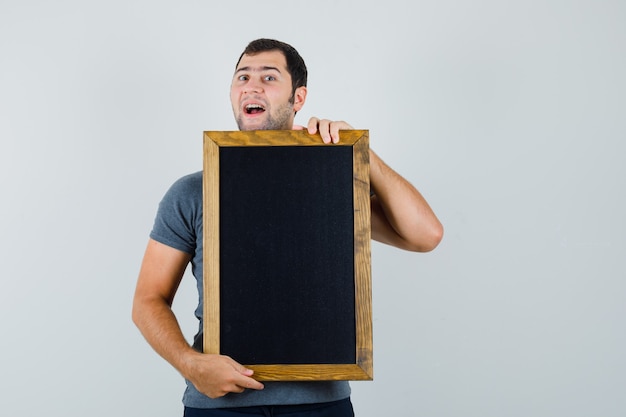 Junger Mann, der Tafel im grauen T-Shirt hält und lustig schaut