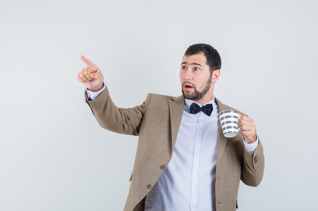 Junger Mann, der Tasse des Getränks hält, während weg im Anzug zeigt und fokussierte Vorderansicht schaut.