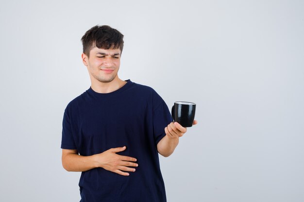 Junger Mann, der Tasse im schwarzen T-Shirt betrachtet und unzufrieden schaut, Vorderansicht.