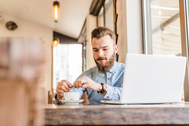 Junger Mann, der Tasse Kaffee mit Laptop auf Schreibtisch hat