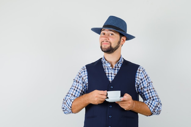 Junger Mann, der Tasse mit Untertasse in Hemd, Weste, Hut hält und zuversichtlich schaut, Vorderansicht.