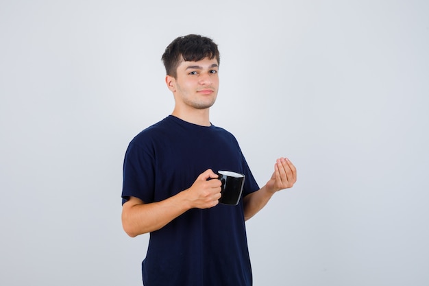 Junger Mann, der Tasse Tee hält, italienische Geste im schwarzen T-Shirt tut und entzückt schaut. Vorderansicht.