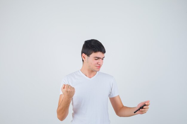 Junger Mann, der Telefon hält, geballte Faust im T-Shirt zeigt und fröhlich aussieht. Vorderansicht.