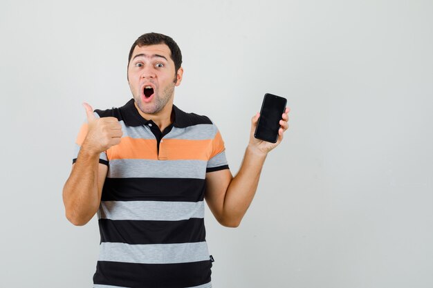 Junger Mann, der Telefon zeigt, während Daumen oben im T-Shirt zeigt und aufgeregt, Vorderansicht schaut.