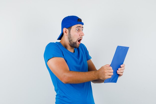 Junger Mann, der über Zwischenablage in blauem T-Shirt und Mütze schaut und schockiert schaut