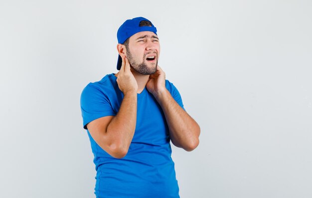 Junger Mann, der unter Nackenschmerzen im blauen T-Shirt und in der Mütze leidet und müde aussieht