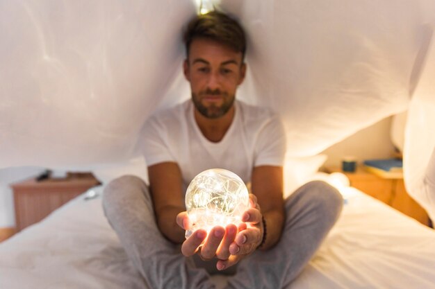 Junger Mann, der unter Vorhang auf dem Bett hält beleuchtete Kugel in der Hand sitzt