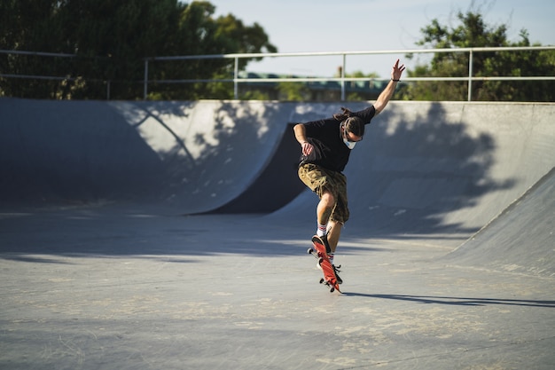 Junger Mann, der verschiedene Tricks mit einem Skateboard im Park tut, der eine medizinische Gesichtsmaske trägt