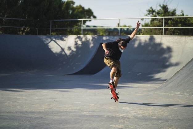 Junger Mann, der verschiedene Tricks mit einem Skateboard im Park tut, der eine medizinische Gesichtsmaske trägt