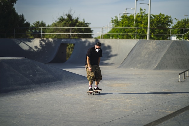 Junger Mann, der verschiedene Tricks mit einem Skateboard im Park tut, der eine medizinische Gesichtsmaske trägt
