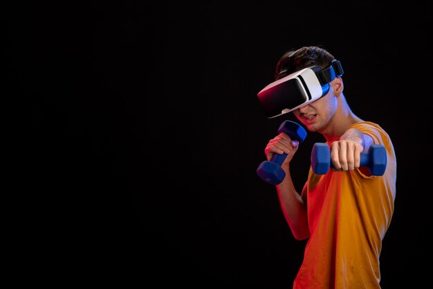 Junger Mann, der virtuelle Realität mit Hanteln auf dunkler Oberfläche spielt