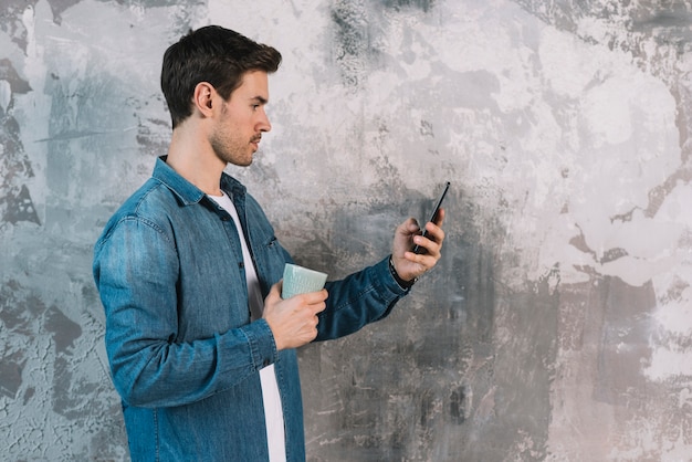 Junger Mann, der vor der verwitterten Wand betrachtet das Mobiltelefon hält Kaffeetasse steht