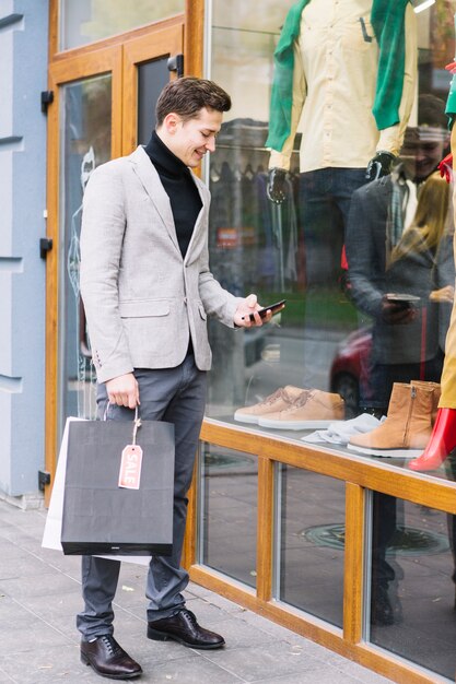 Junger Mann, der vor Shop unter Verwendung des Smartphone steht