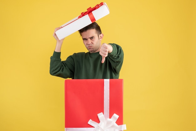 Junger Mann der Vorderansicht, der Kastenabdeckung hält, das Schild unten macht, das hinter großer Geschenkbox auf Gelb steht