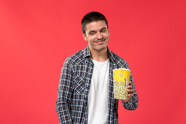 Junger Mann der Vorderansicht, der Popcorn-Paket hält und leicht auf hellrotem Wandkino-Filmfilm lächelt