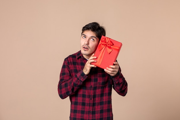 Junger Mann der Vorderansicht, der rotes Paket mit Geschenk für Frauen an brauner Wand hält