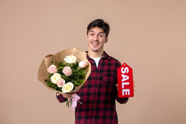 Junger Mann der Vorderansicht, der schöne Blumen und Verkaufsnamensschild an brauner Wand hält