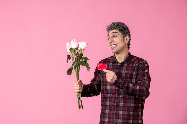 Junger Mann der Vorderansicht, der schöne rosa Rosen und Bankkarte an rosa Wand hält