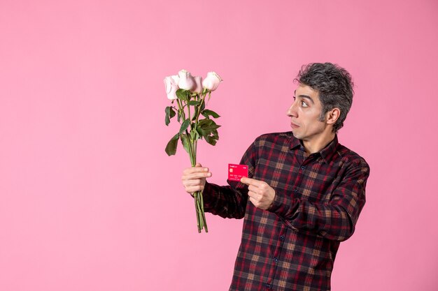 Junger Mann der Vorderansicht, der schöne rosa Rosen und Bankkarte an rosa Wand hält