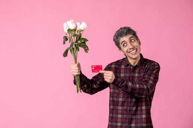 Junger Mann der Vorderansicht, der schöne rosa Rosen und Bankkarte an rosa Wand hält