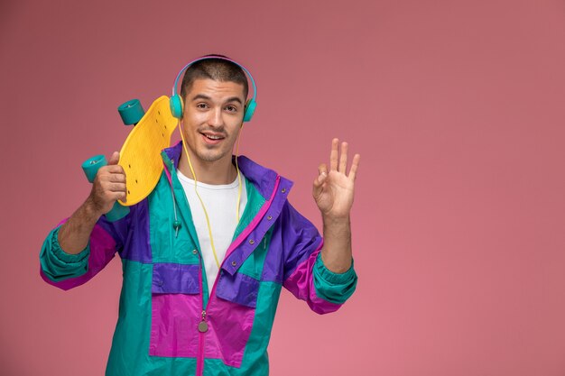 Junger Mann der Vorderansicht im bunten Mantel, der Musik hört, die Skateboard auf rosa Schreibtisch hält