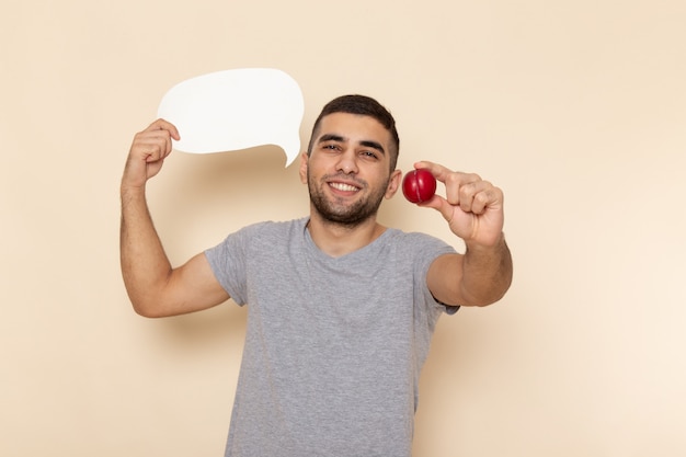 Kostenloses Foto junger mann der vorderansicht im grauen t-shirt und in den blauen jeans, die weißes zeichen und pflaume auf beige halten