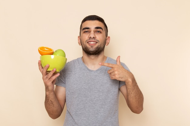 Junger Mann der Vorderansicht im grauen T-Shirt und in den blauen Jeans lächelnd und hält Platte mit Früchten auf Beige