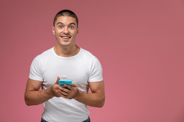 Junger Mann der Vorderansicht im weißen Hemd unter Verwendung seines Telefons und Lächeln auf rosa Hintergrund