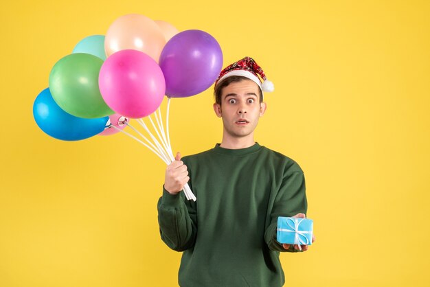 Junger Mann der Vorderansicht mit großen Augen mit Weihnachtsmütze und bunten Luftballons, die blaue Geschenkbox auf Gelb halten