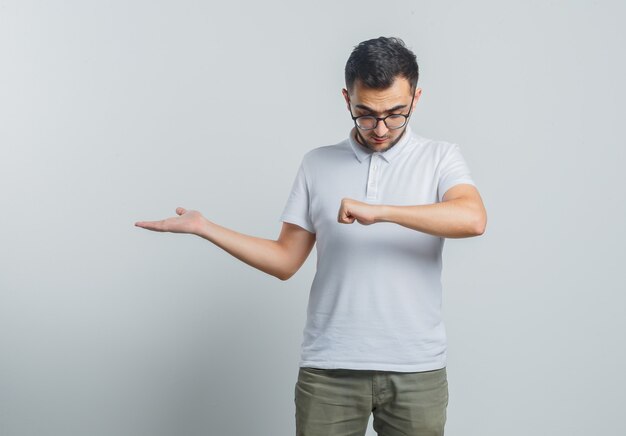 Junger Mann, der vorgibt, auf die Uhr am Handgelenk zu schauen, etwas im weißen T-Shirt, in der Hose zeigt und konzentriert schaut